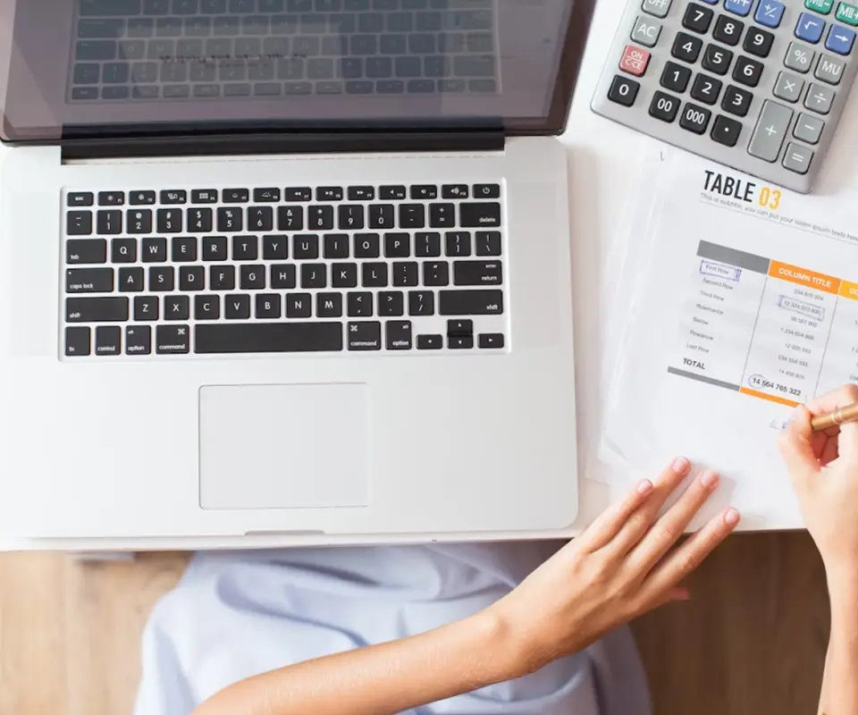 A woman writing on a paper, and on the table a laptop and a calculator.