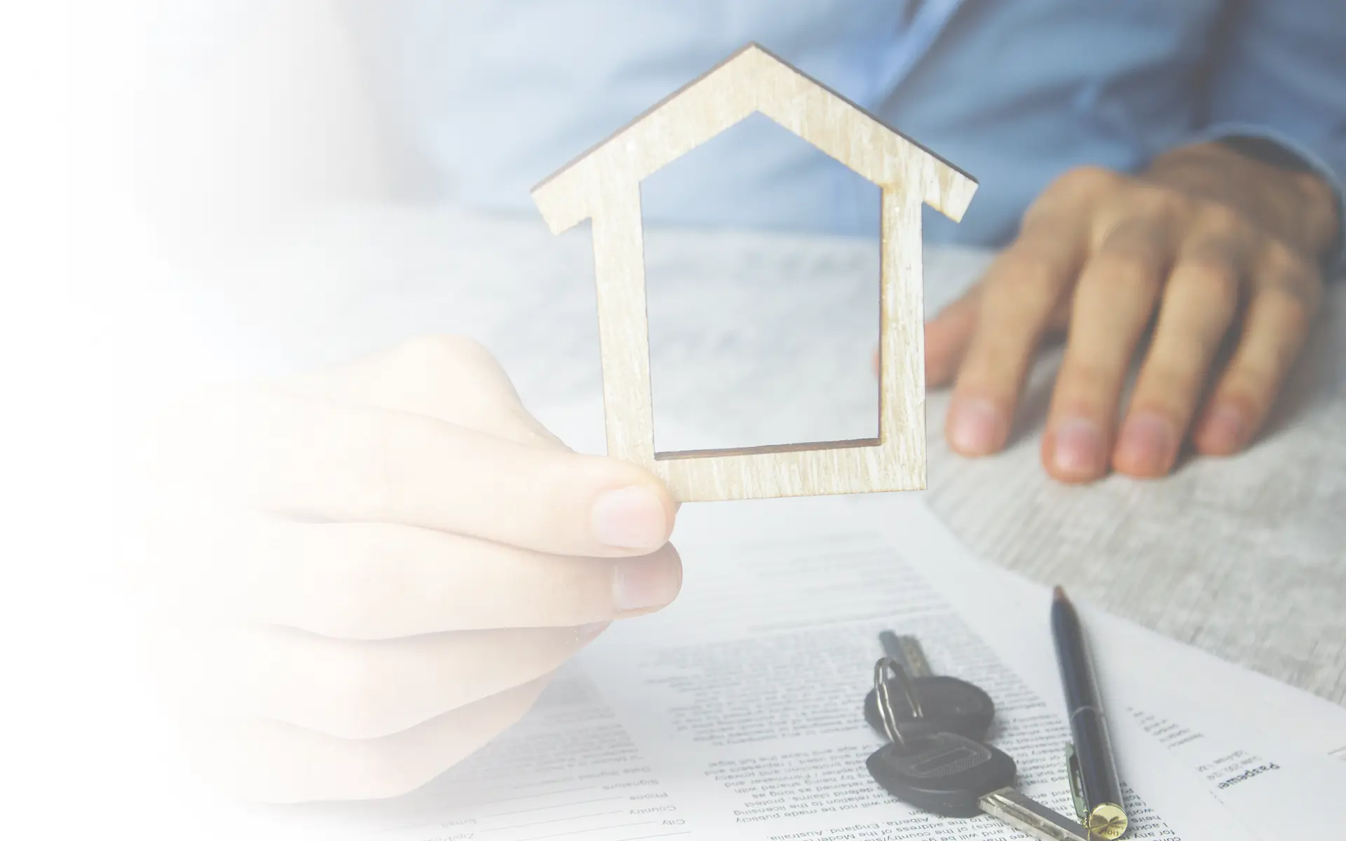 A man is holding a wooden house shape, and on the table, there are some keys and a pen.